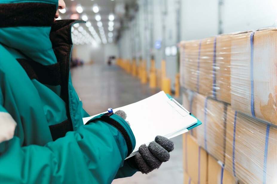 A worker in a teal insulated jacket and gloves holds a clipboard while inspecting shrink-wrapped boxes inside a cold storage warehouse.